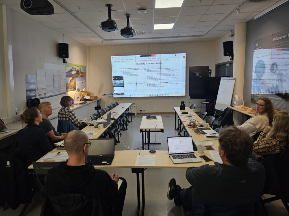 Et møte med en gruppe mennesker rundt et bord og med en presentasjon på skjermen