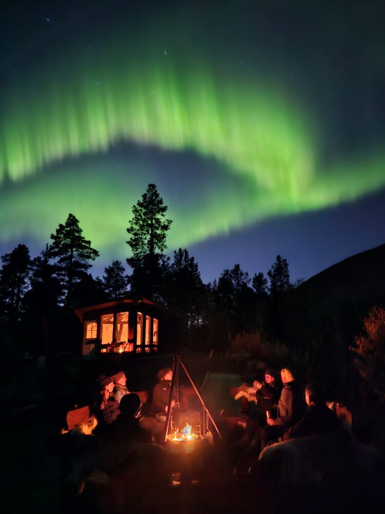En gruppe mennesker rundt bålet med hytte og nordlys i bakgrunnen