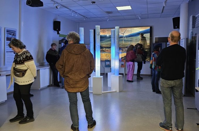 Flere personer beveger seg rundt i et moderne utstillingslokale med hvite vegger og takbelysning. I midten står en installasjon med fem høye, sylindriske rør som lyser svakt blått, montert på en hvit sokkel. I bakgrunnen vises et stort landskapsbilde med vindturbiner på en veggskjerm. Noen personer står og ser på utstillingene eller snakker sammen, mens andre går gjennom rommet. Informasjonstavler og mindre utstillingselementer er plassert langs veggene.