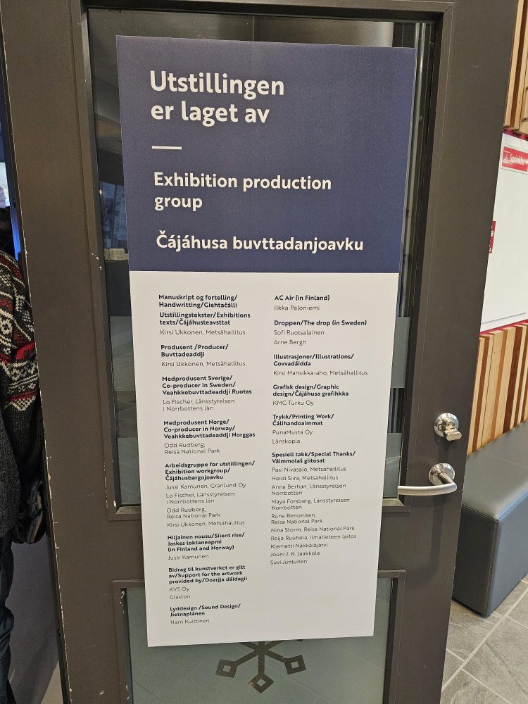 En plakat montert på en dør, med overskriften «Utstillingen er laget av / Exhibition production group Čáhces buvttadanjoavku». Under overskriften står en liste over bidragsytere, produsenter og samarbeidspartnere på flere språk. Plakaten er todelt, med et mørkeblått felt med overskrift øverst og et hvitt felt med tekst nederst.