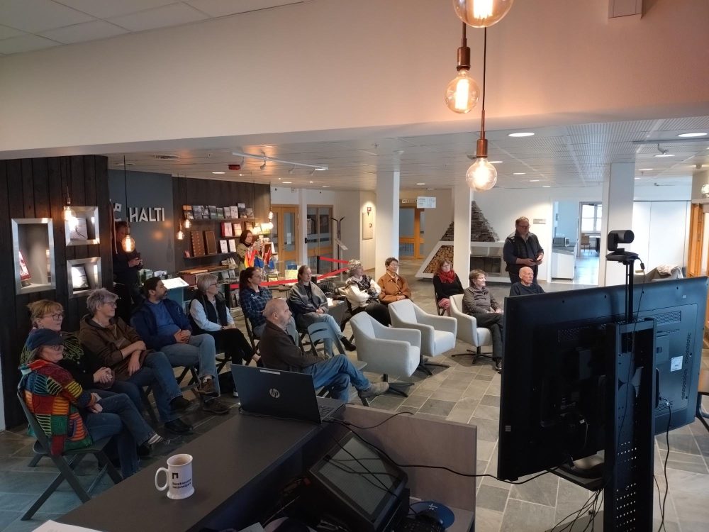 Publikum sitter på stoler i et lyst innendørs lokale og ser mot en presentasjon som vises på skjermer foran dem. I forgrunnen står en skjerm vendt mot publikum. Bak publikum er det en vegg med bøker og utstilling, der teksten «HALTI» er synlig. Enkelte personer står bakerst i rommet. Hengende lyspærer gir varm belysning.