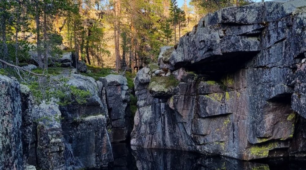 Smalt juv med bratte fjellvegger og stille vann nederst, omgitt av furuskog og store steinblokker i et solbelyst landskap.