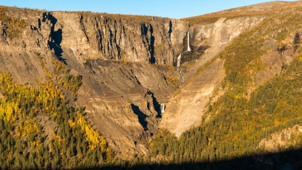 Bratt fjellvegg med et smalt fossefall som renner ned i en steinete kløft, omgitt av høstfarget skog i gul- og grønntoner under klar himmel.