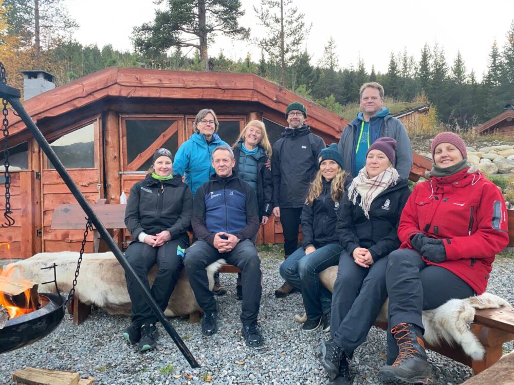Gruppe mennesker som sitter og står med hytte i bakgrunnen