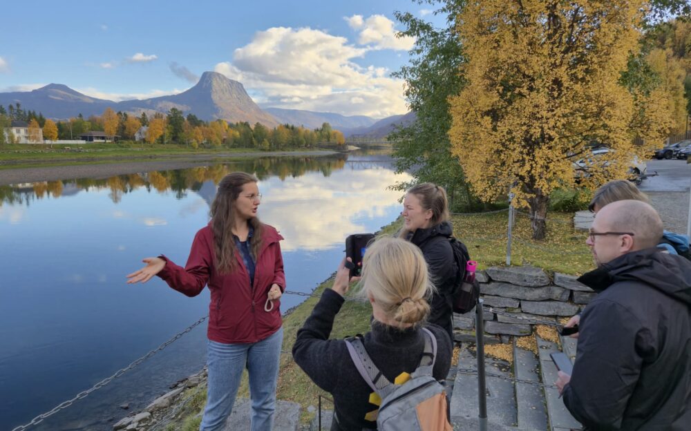 Gruppe med mennesker der en forklarer og en tar bilder med elv og høst lanskap i bakgrunnen